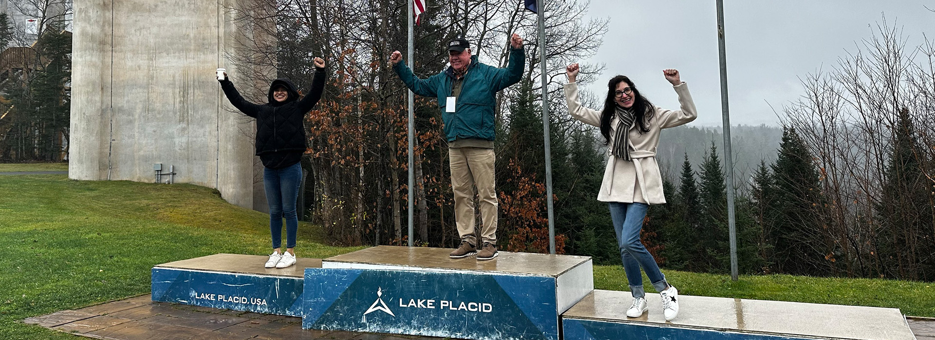 Three Lake Placid Film Festival participants stand smiling with arms raised triumphantly on an Olympic-style podium outside at the base of the ski jumps.
