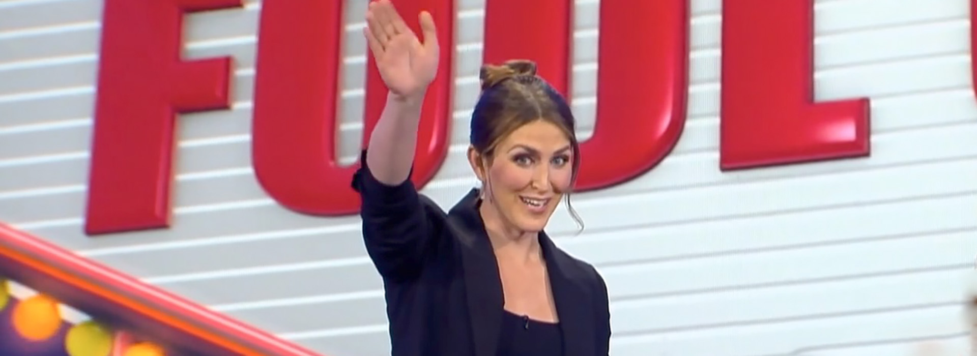 Performer Alyx Hilshey on stage, waving at her audience with red letters on a white background behind her.