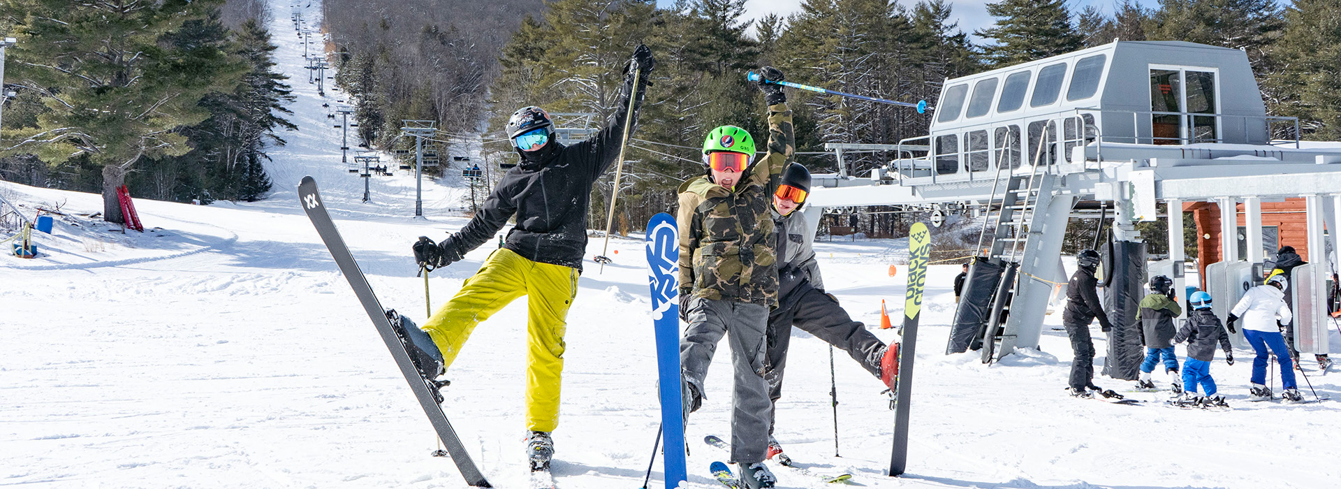 Three young skiers stand with one ski raised in front of the old Hudson Chair, celebrating a bright beautiful day.