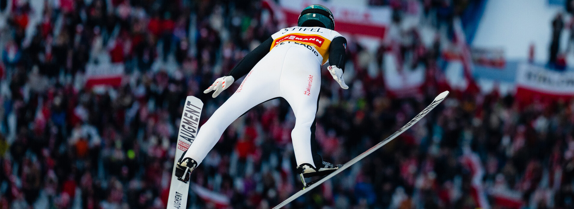 An elite ski jumper in a white unitard soars through the air high above a big crowd of spectators below.