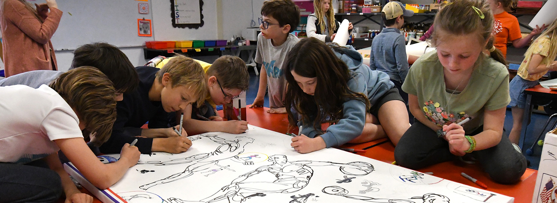 Seven kids in a classroom surround a black and white line art drawing as they sketch and color the art together.