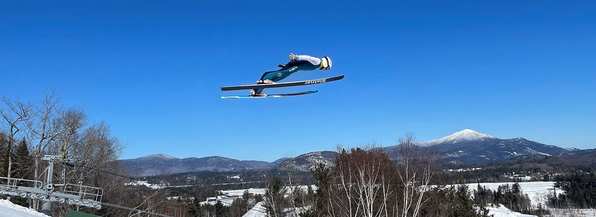 Tate Frantz soaring off the big jump at Lake Placid against a blue sky with Whiteface Mountain in the background.