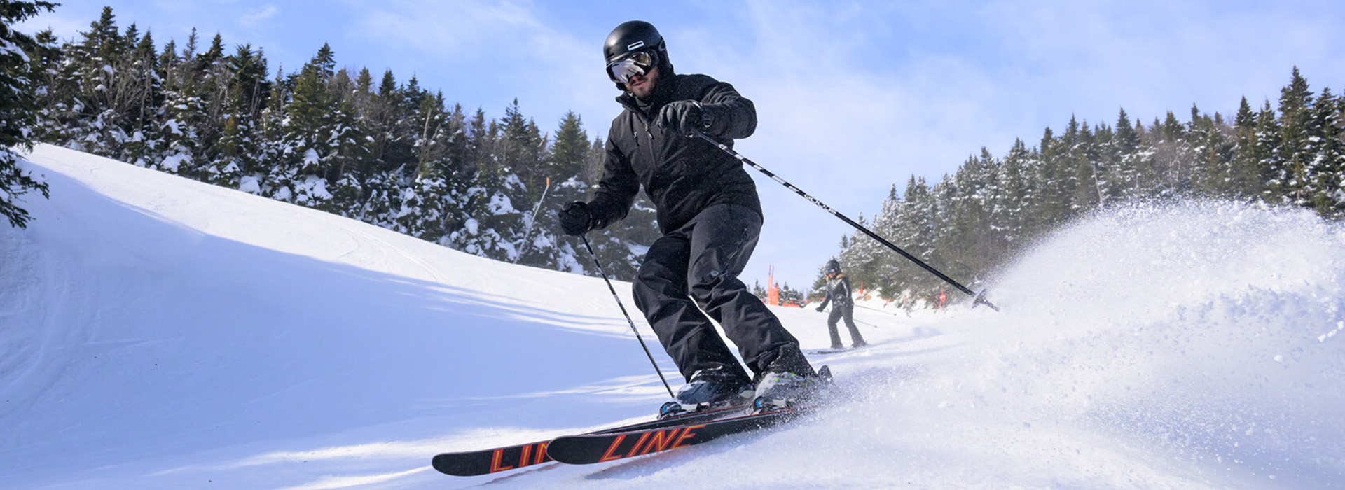 A male skier in black makes a turn just in front of the camera on a steep slope with trees and another skier in the background.