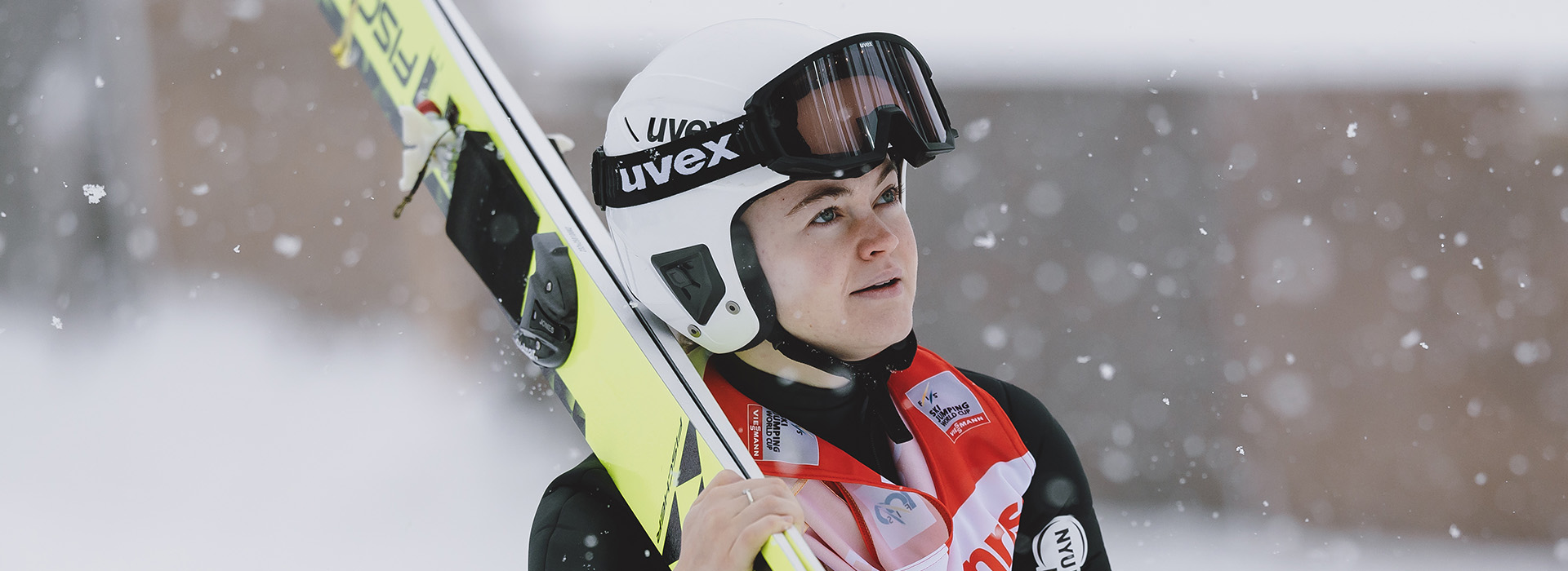 A female ski jumper with skis over her shoulder and helmet on looking upward and forward with snow falling gently in the background.
