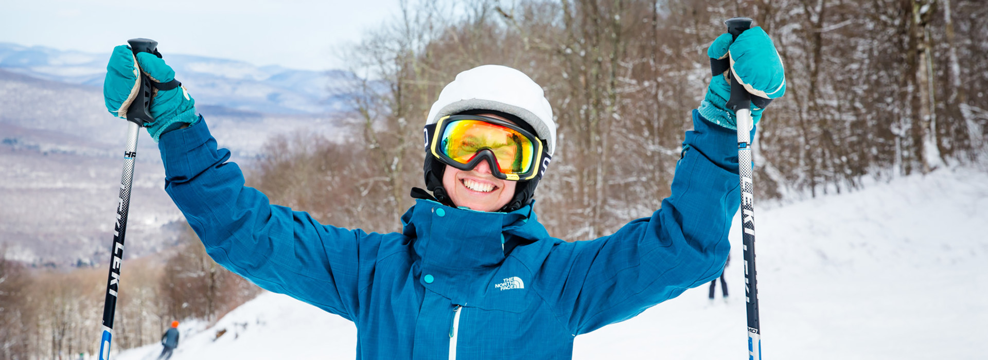 A happy, smiling female skier in a blue jacket and white helmet raises her arms and poles in joy on top of Belleayre.