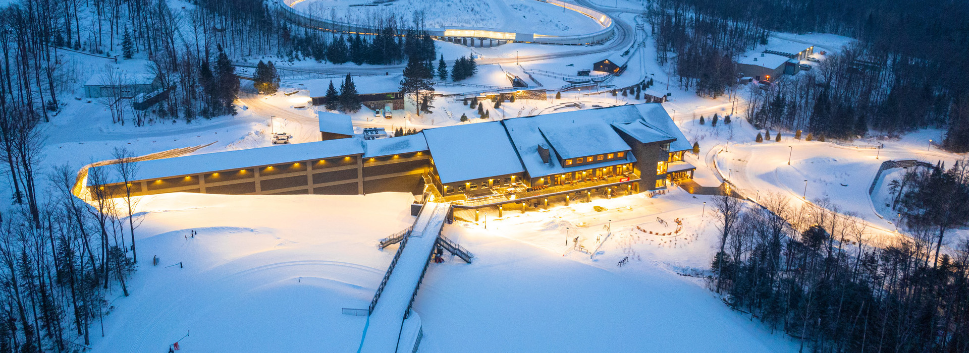 An aerial view of the Mountain Pass Lodge at Mt Van Hoevenberg in winter early in the morning with lights on the lodge in the low lights of the dawn.
