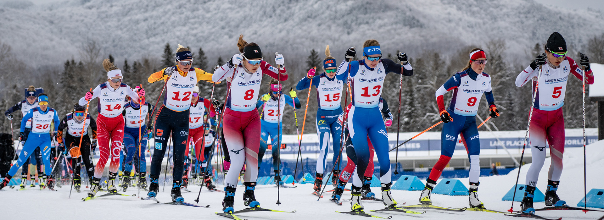 A large group of women cross country ski racers in a mass start with snowy mountains in the background.