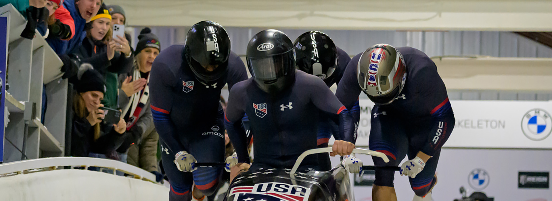 A four man Team USA bobsled just starting down the track with spectators yelling.