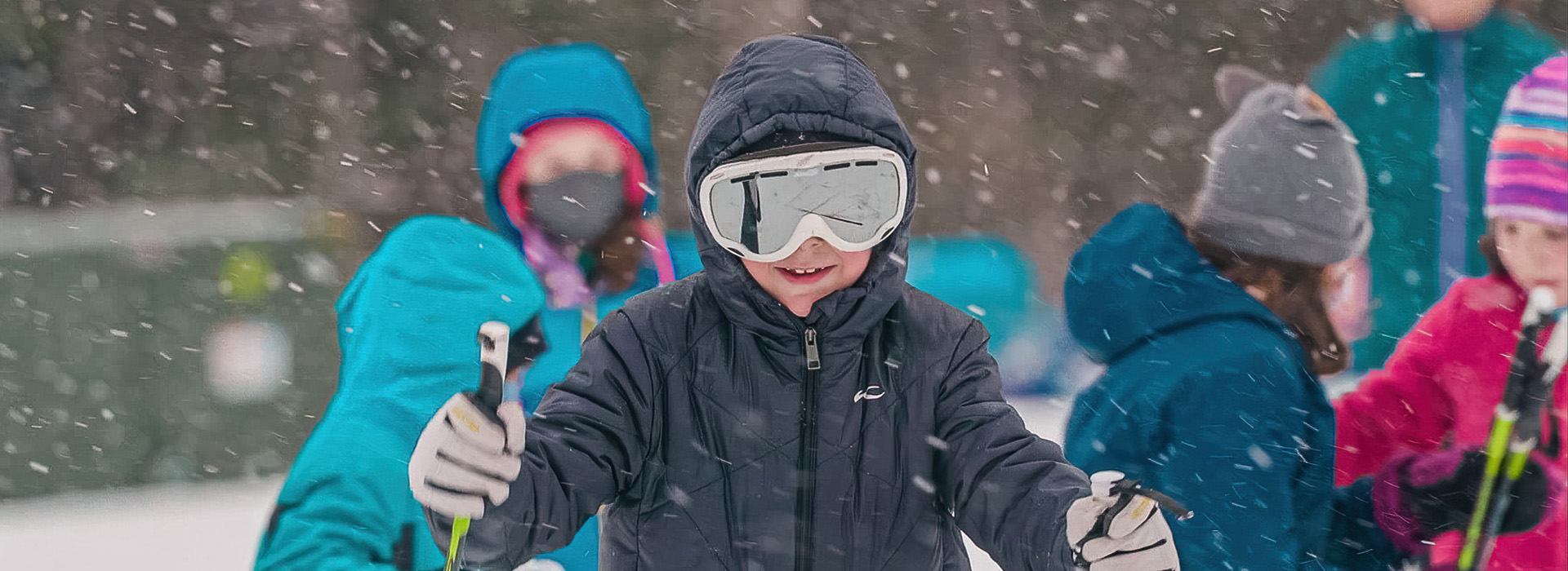 A youth cross country skier with goggles looks at the camera and smiles, with snow flying around him and other children in the background.