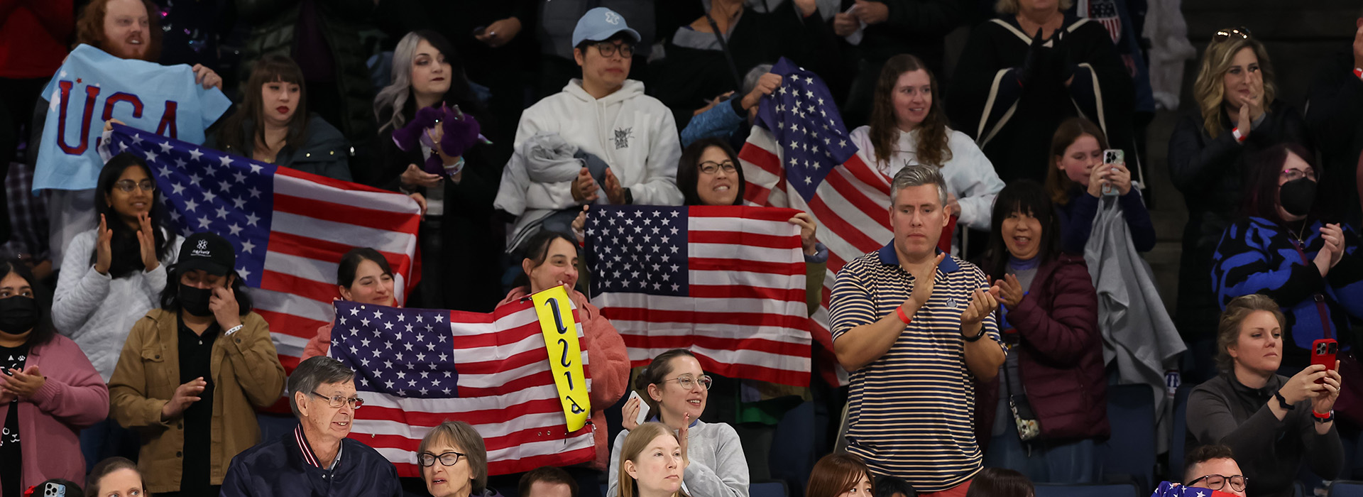 Part of a large crowd of spectators at a Skate America competition, all holding flags and banners and clapping.