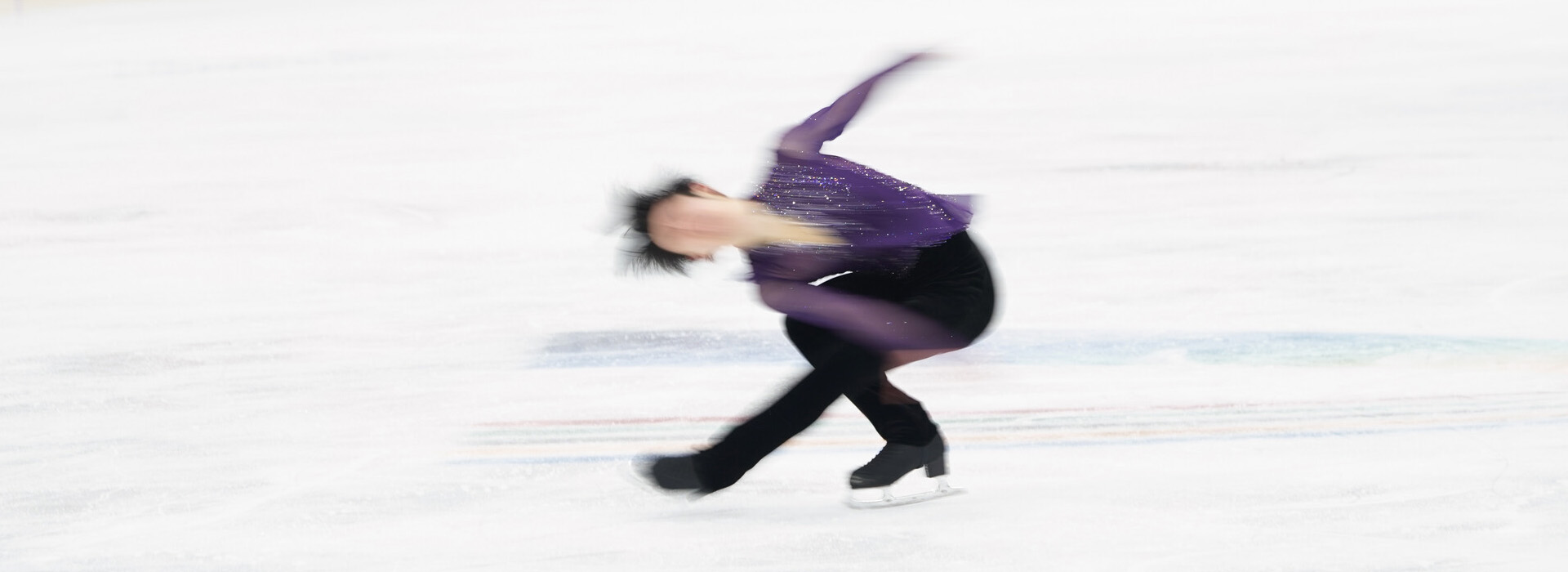 A male figure skater in a purple top and black pants shown in a blur as he spins on the ice.
