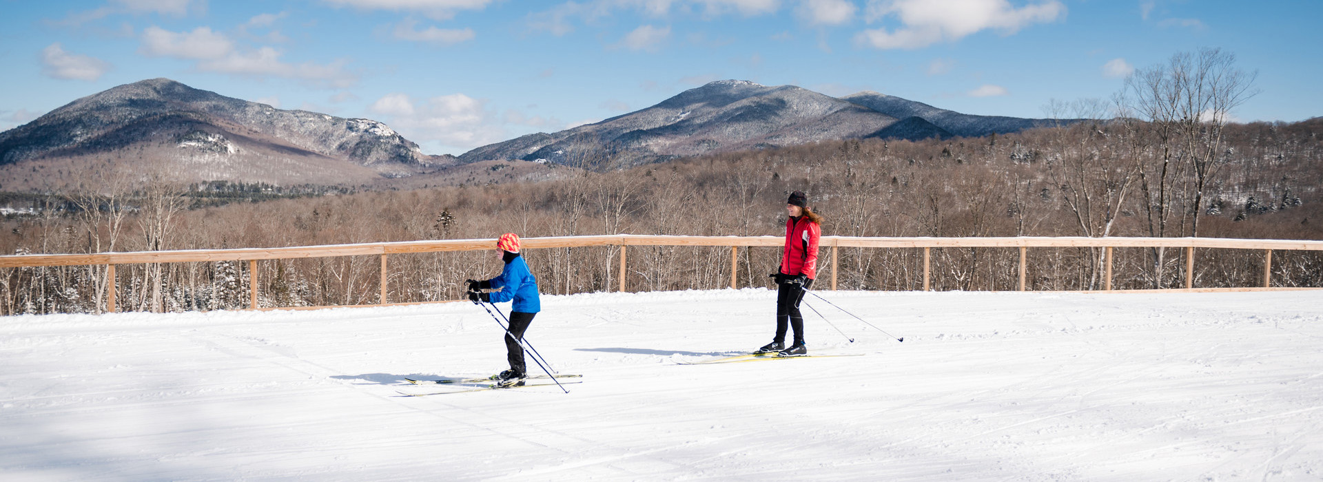 An adult in a red jacket follows a young skier in a blue jacket across a wide Nordic ski trail at Mt Van Hoevenberg with the mountains behind them in the background.