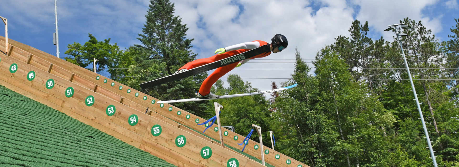 On a beautiful summer day, a ski jumper in an orange unitard sails through the air just above the green grassy landing area