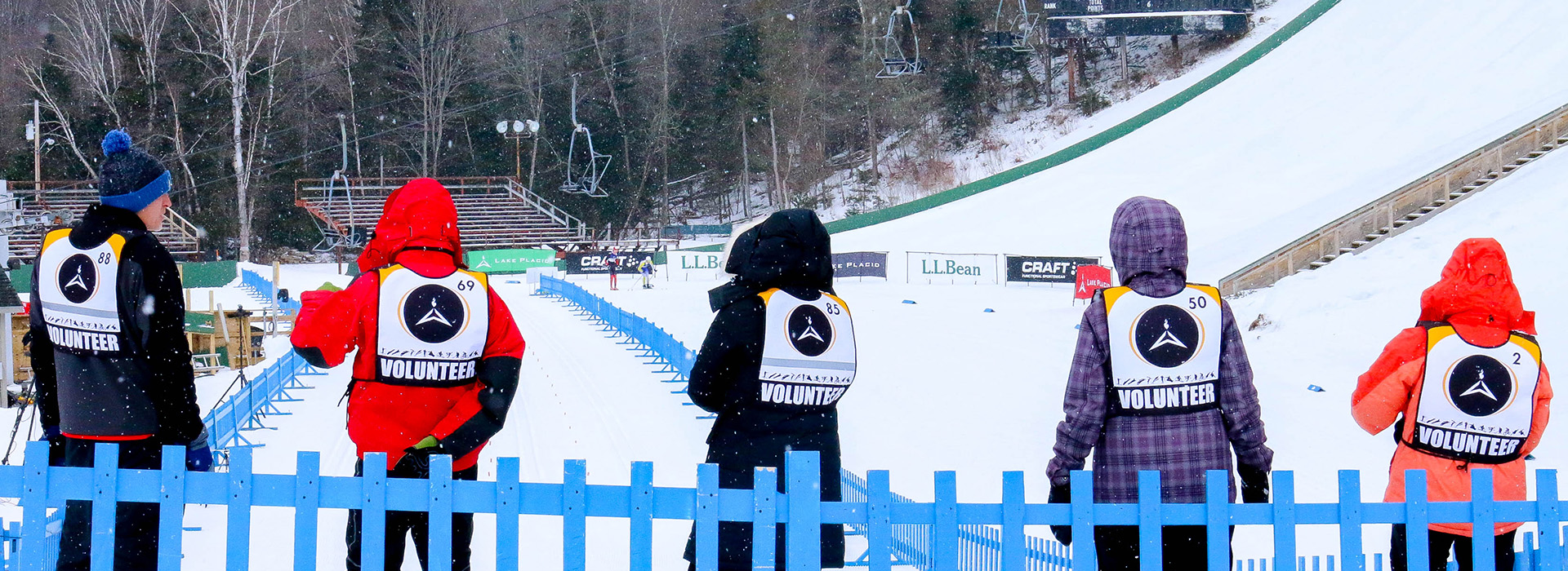 Five people with volunteer bibs stand facing away from the camera on a winter day at the base of the big jumps at the Olympic Jumping Complex.