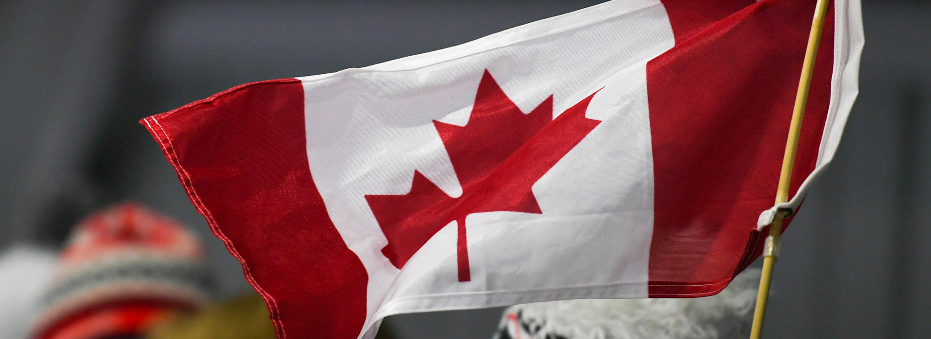 A handheld Canadian flag is the central point of this image with an out of focus spectator hat in the background.