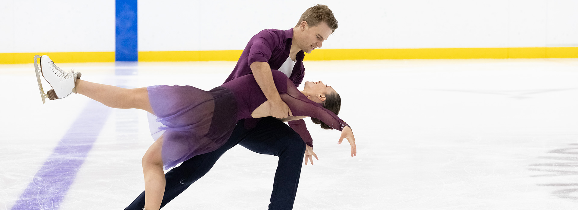 Ice dancers in purple outfits on the ice in the 1980 Rink with the male skater bending the female skater over backwards as they move across the rink.