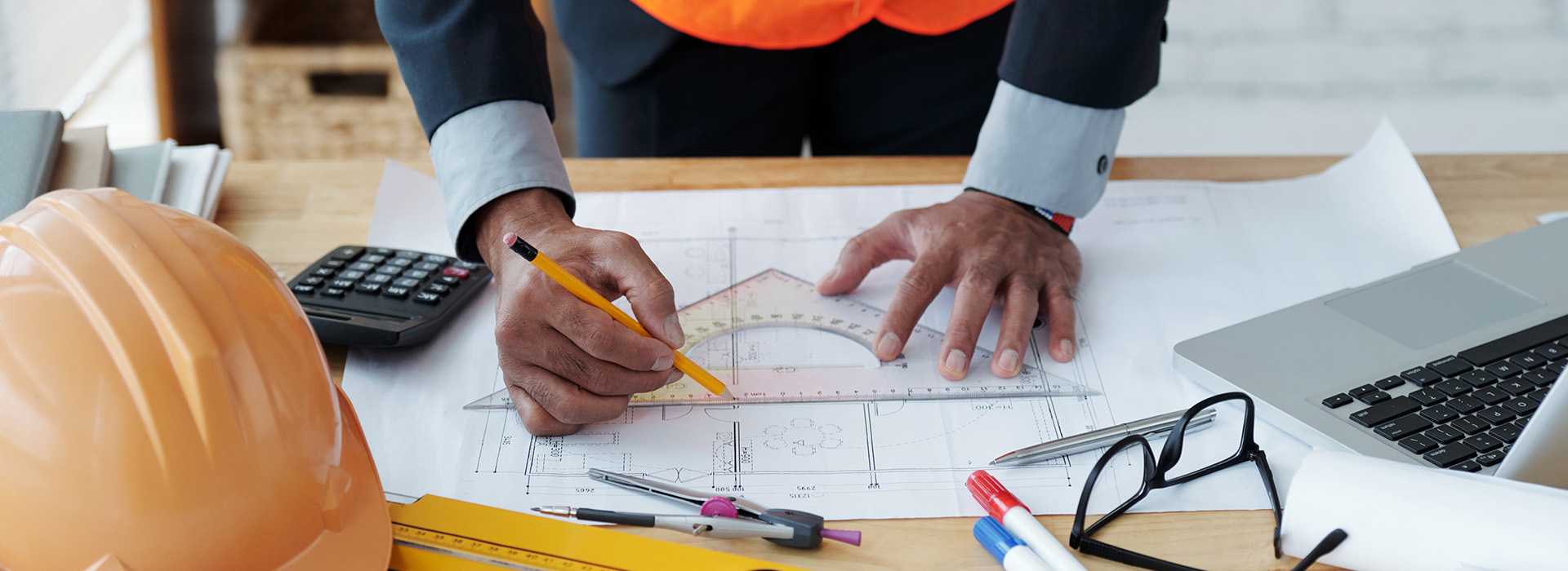 A professional's hands with a triangle and pencil mark up architectural drawings on a table with a hardhat and other implements alongside the drawing.