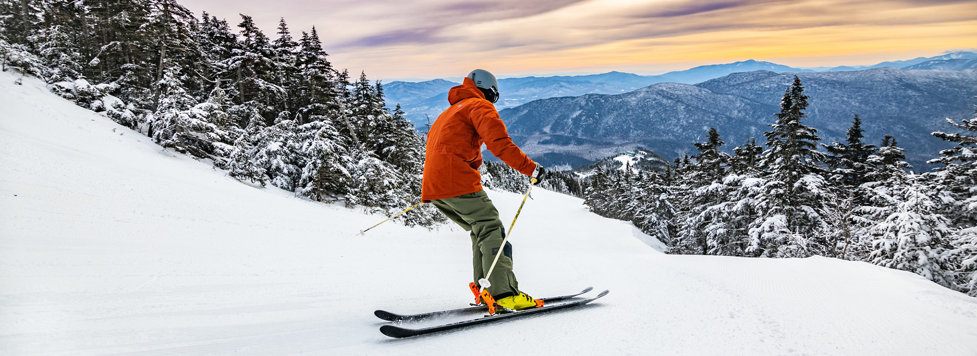 Skier in an orange jacket descends the snow covered slopes of Whiteface high on the mountain with the rising sun and trees and mountains in the background.