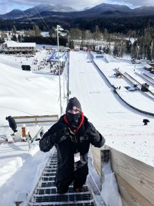 Asa Mikelbank standing with thumbs up on the stairs on the side of the big jump hill while wearing heavy winter gear, including a hat and facemask. 