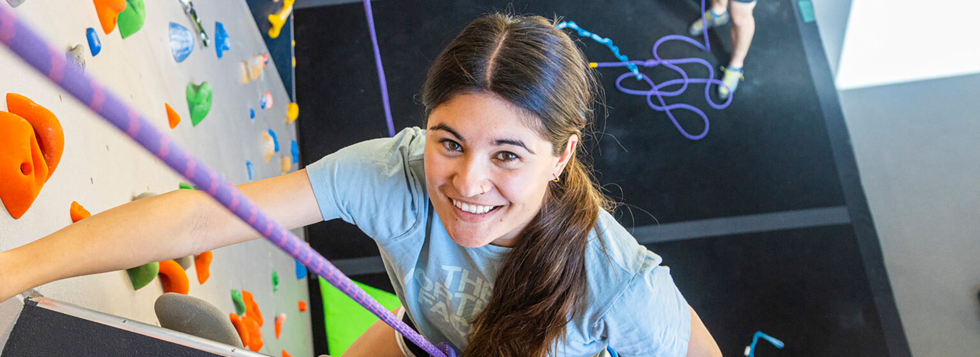 A young female climbing up the Hi Notch wall, smiling into the camera above her with hand holds and rope and the floor around her and in the background.