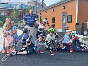 Skating coach Tom Miller standing with a group of youth rollerblade skaters surrounding him, some sitting, lying, kneeling, etc, all happy to be there with one exhaulting Tom with her hands in the air as if to say, "how great is he?" 