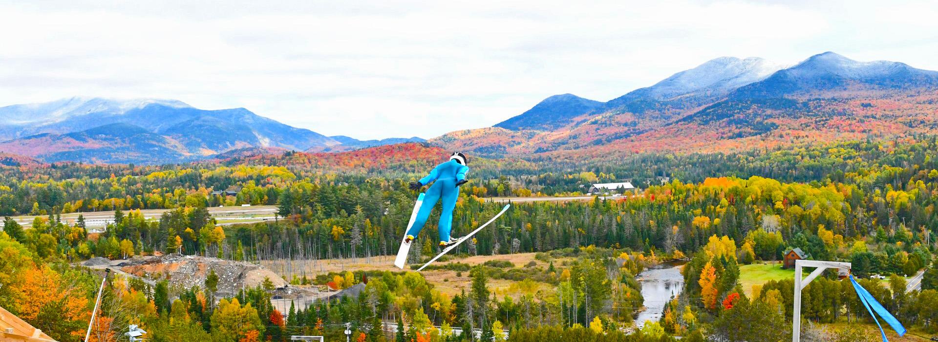 A ski jumper in a blue suit sails through the air over a colorful fall landscape with mountains in the background.