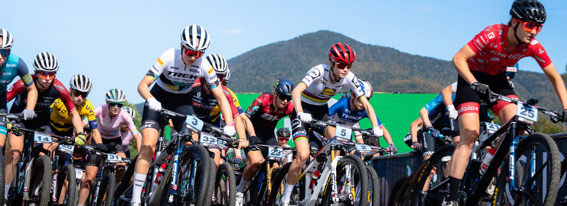 A mass start of a women's UCI mountain bike race close up with many riders and a mountain and blue sky in the background.
