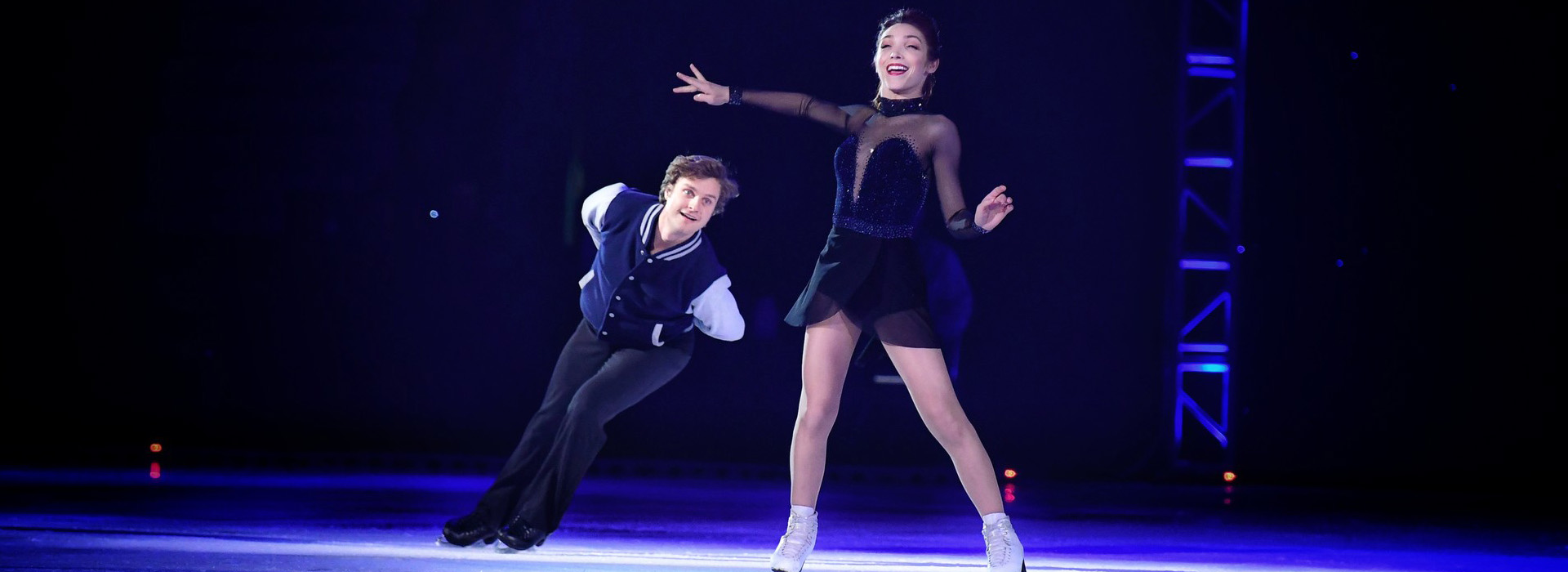 A female figure skater smiles brightly as her male pairs counterpart skates around her and under her outstreached arm.