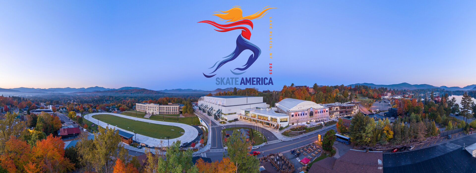An aerial photo of the Lake Placid Olympic Center on a fall day with a blue sky and the Skate America logo above the Olympic Center.