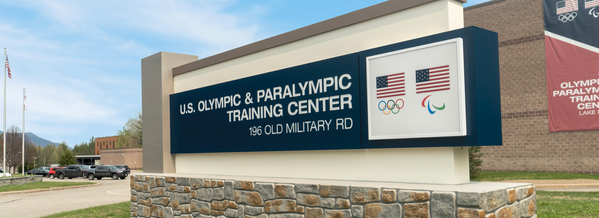 Image of the large sign for the U.S. Olympic & Paralympic Training Center, 196 Old Military Road with two american flags and Olympic symbols.