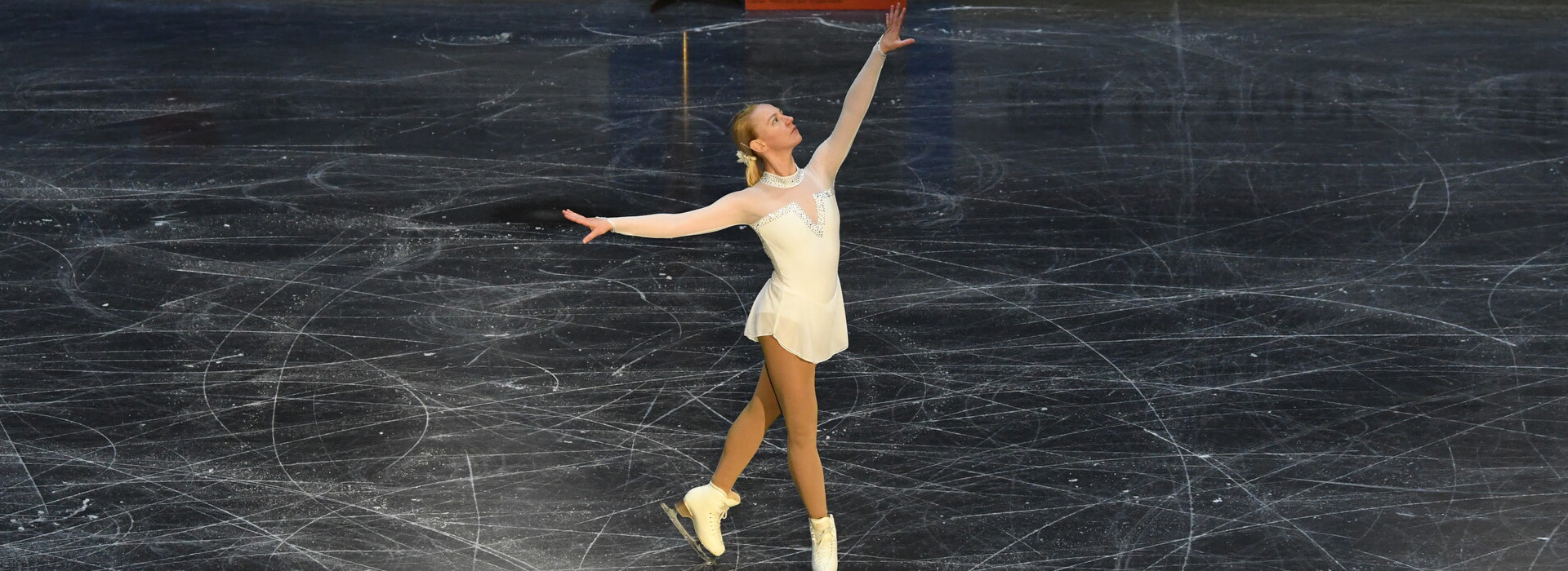 A female skater in a white outfit stops on the black ice surface with her arms raised in an artistic fashion.