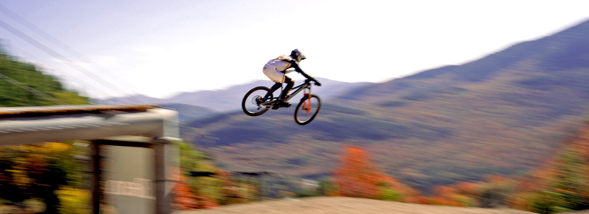 An elite mountain bike rider sails through the air high off a jump at Whiteface Mountain with fall foliage and mountains in the background.