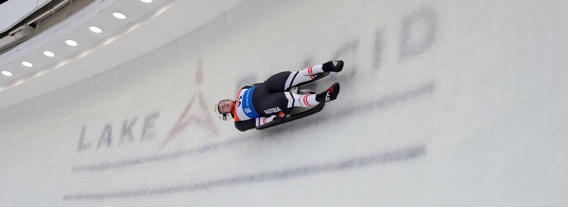 A male luge athlete slides down the track across a big tall turn with the words "Lake Placid" visible in the ice below.