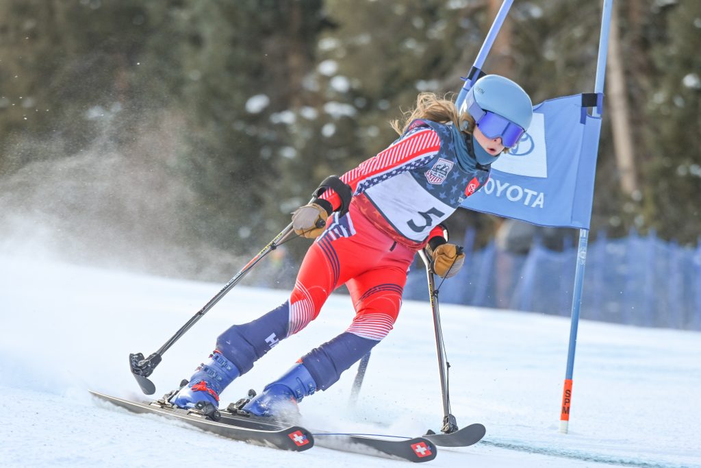 Kelsey, in a red ski suit, skis hard around a race gate with trees in the background.