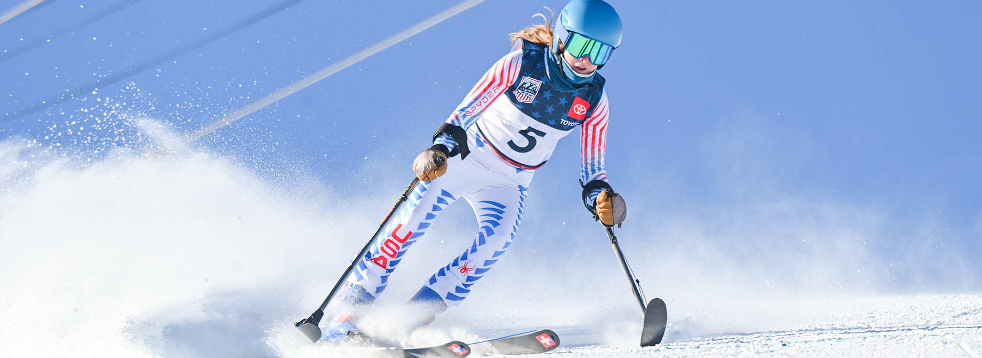 Kelsey O'Driscoll skis with outriggers wearing a white Team USA uniform, all against a blue sky.