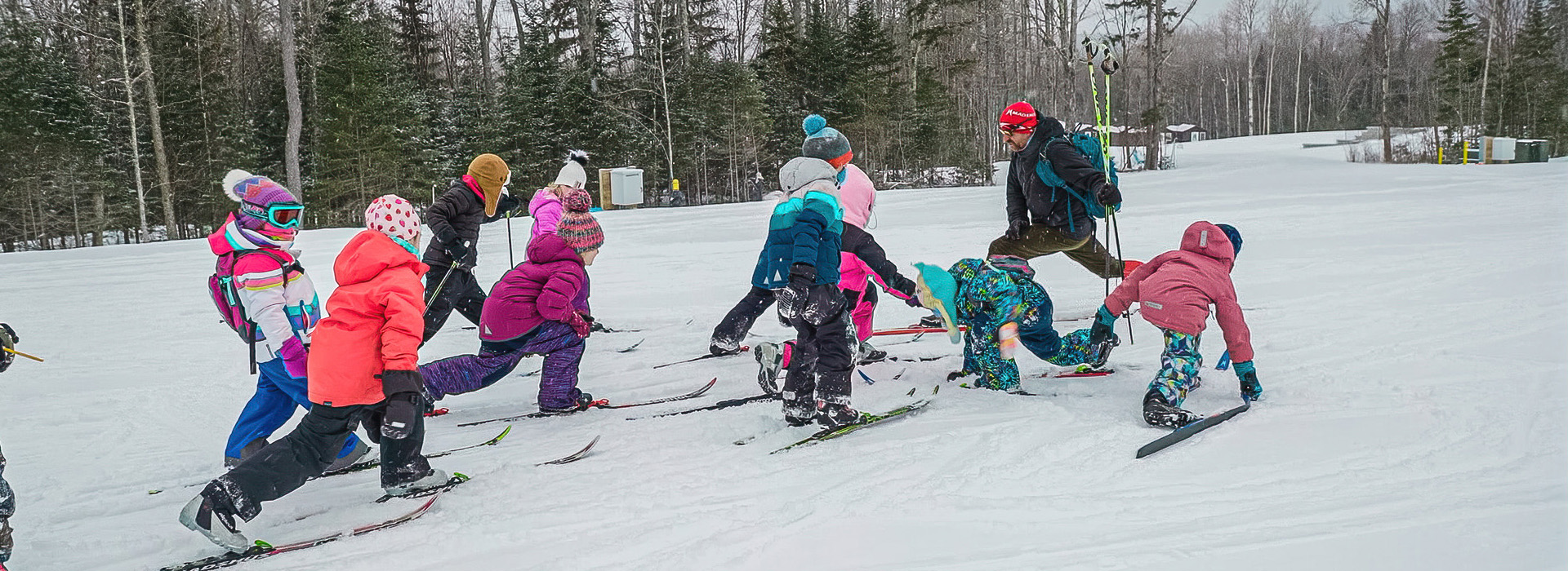 A group of young skiers dressed in winter gear practicing a move on skis their instructor is demonstrating for them.