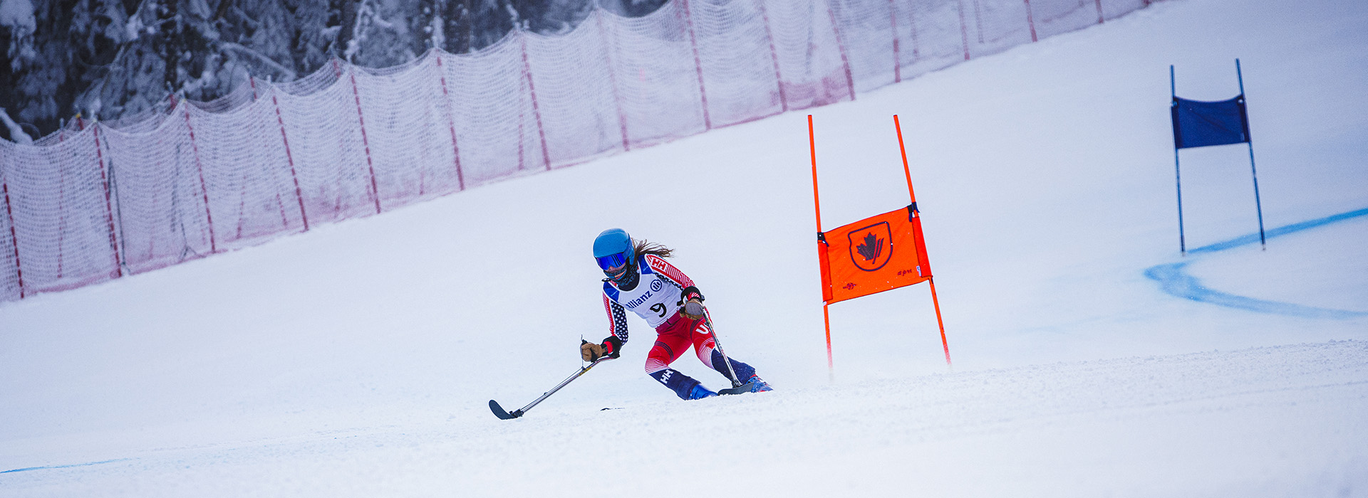 Kelsey, in a red ski racing leotard, speeds down a slope, passing an orange gate with safety fencing and trees in the background.