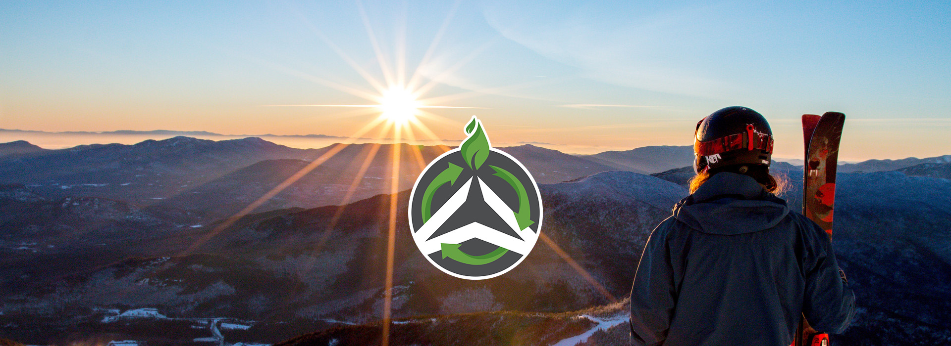 A winter sunrise view from the summit of Whiteface with a skier on the right and the green and white Olympic Authority sustainability logo in the center with the sun shining on it.