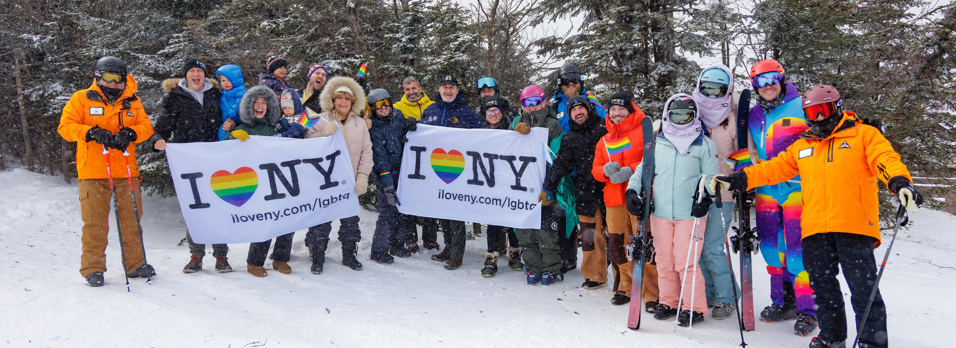 A large group of skiers on the mountain in the snow with trees in the background, some of them holding two white banners with the I Love NY logo, including a rainbow heart in each.