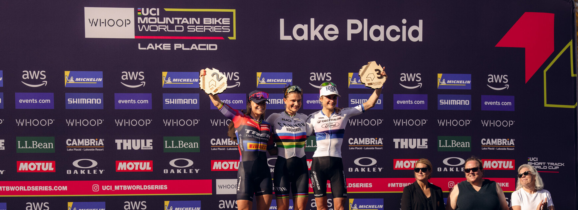 Three women stand triumphantly and holding trophies on the podium after their race at Mt Van Hoevenberg in Lake Placid with the UCI World Series and Lake Placid logos in the background.