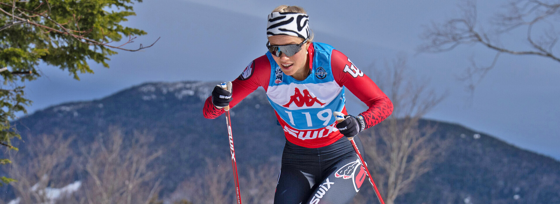 A female cross-country ski racer works hard climbing a trail at Mt Van Hoevenberg with the mountains visible in the background.