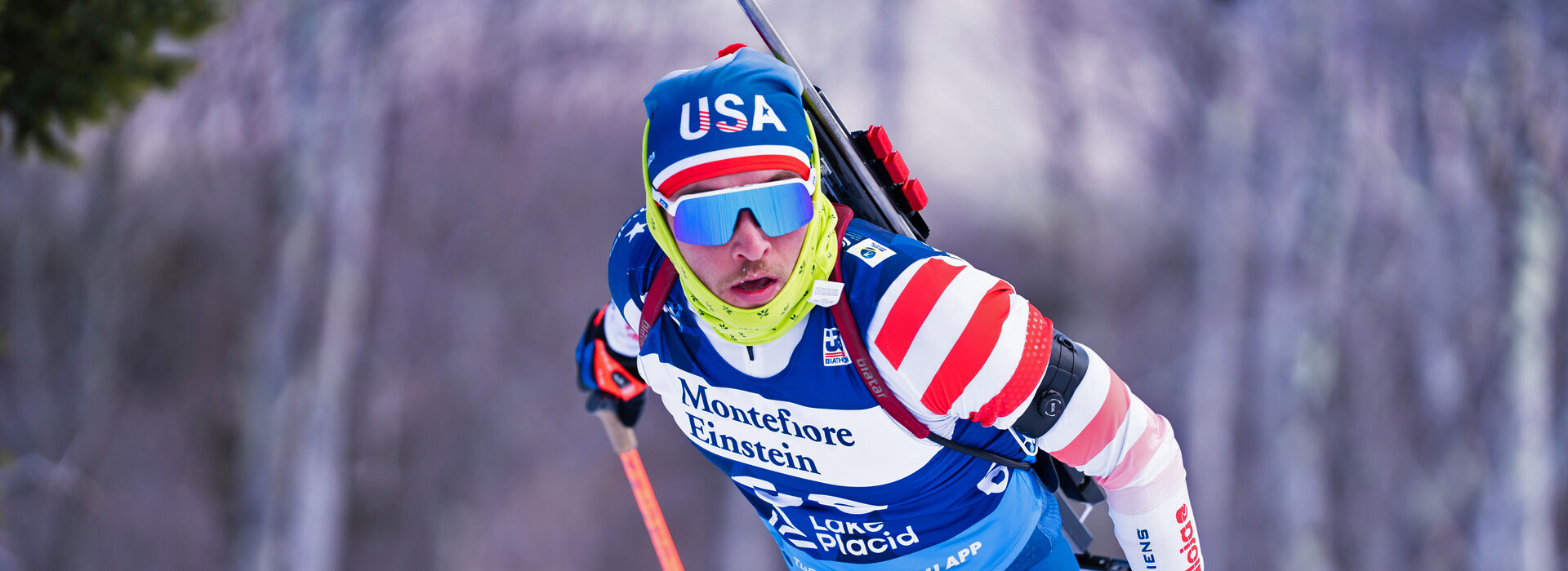 An IBU Cup athlete bearing a USA hat, looks hard through his sunglasses as he works hard skiing in competition on a winter day.