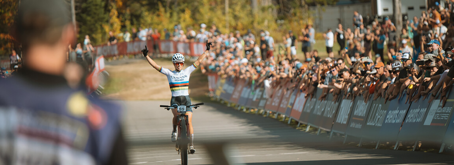 A female rider in a UCI mountain bike cross country race raises her arms in triumph as he crosses the finish line in front of many spectators in an outdoor fall scene.