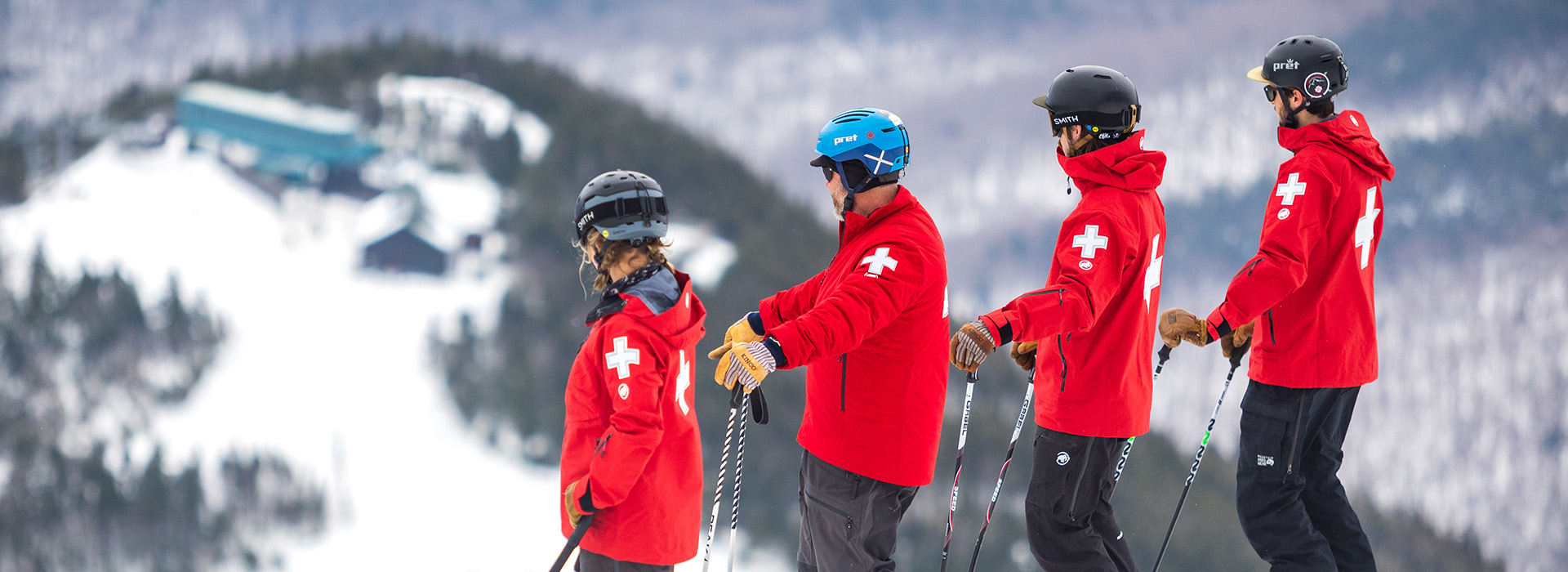 Four ski patrol members of the Whiteface Mountain crew, all in red Mammut jackets on the summit looking down on Little Wihteface.