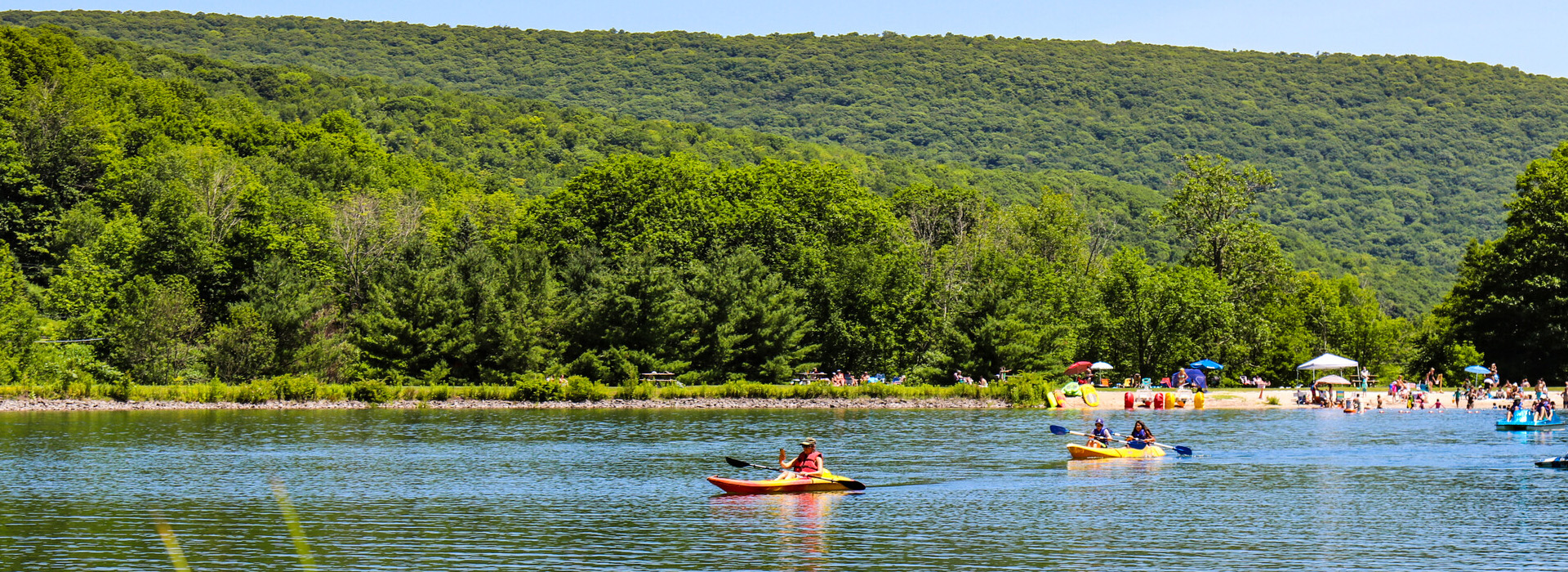 A kayaker floats across the lake at Belleayre Beach on a bright summer day with the green forest of the mountain in the background.
