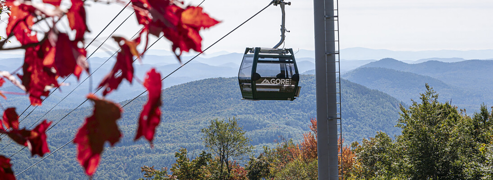 A cabin of the Northwoods Gondola rises high on Gore Mountain with fall foliage in the foreground and mountains in the background.