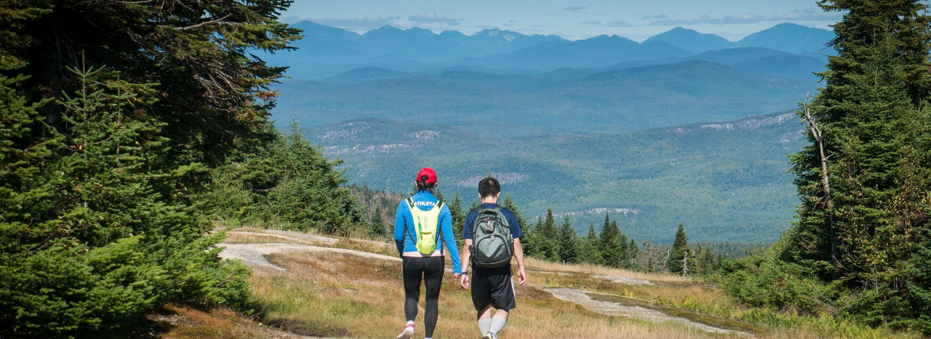 Two hikers high on Gore Mountain in the summer walking away from the camera toward the background view of the mountains.