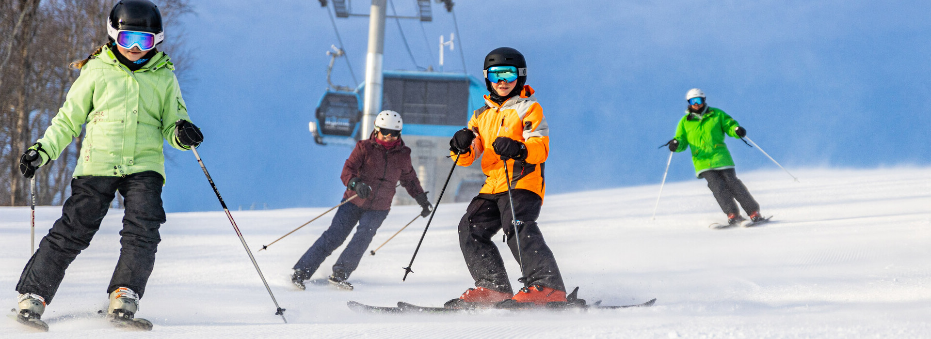 A family of four skis down a slope facing the camera with the gondola overhead and blue sky behind them.