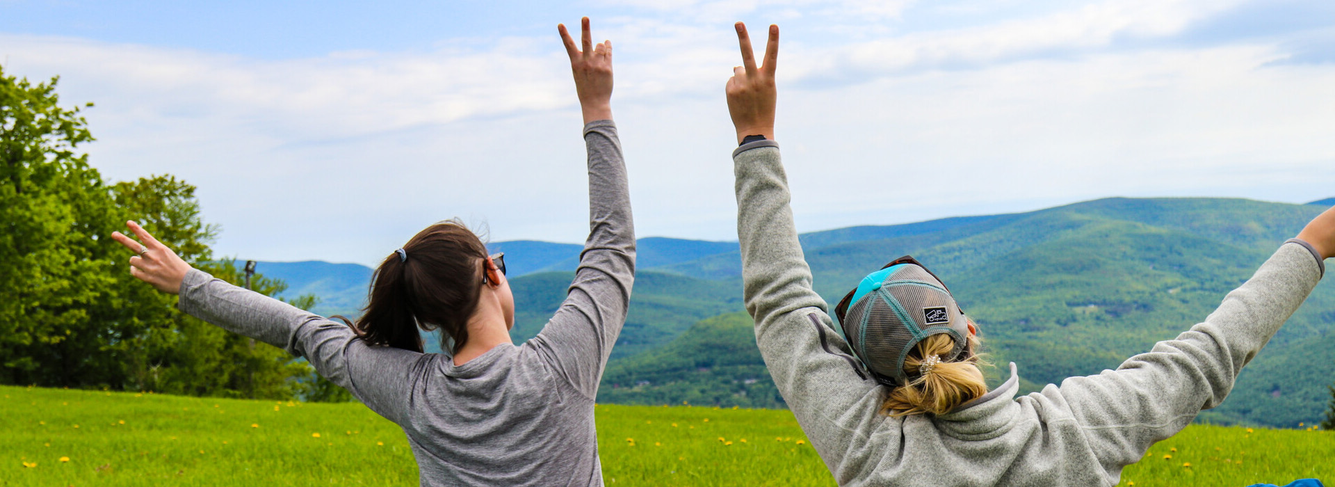 Two women facing away from the camera and sitting in the grass on the Belleayre Mountain summit raise their hands in peace signs in a signal of happiness.