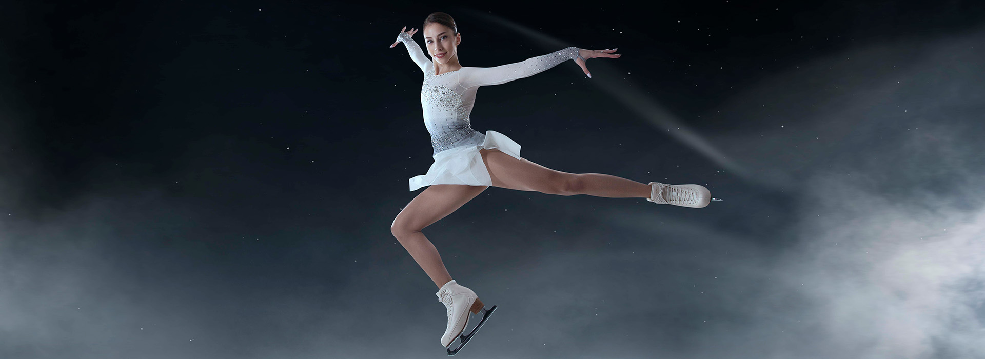 A young female figures skater in a white uniform jumping with her hands and legs outstreatched in an artful pose against a black background with a few with wispy cloud-like features.